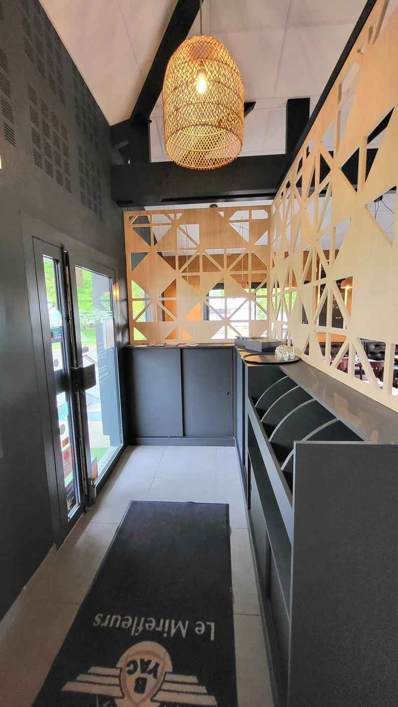 Entrée de restaurant avec claustras géométriques en bois, comptoir sombre et suspension en fibres naturelles