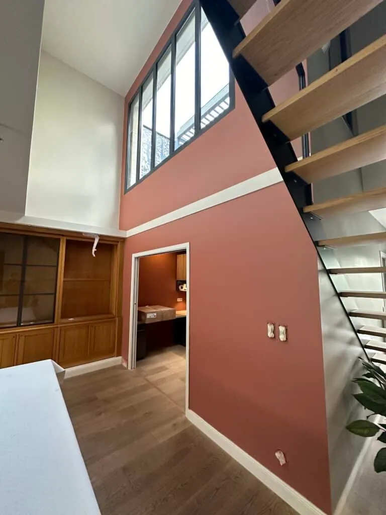 Intérieur rénové avec mur rose poudré, escalier en bois et métal, verrière haute et bibliothèque en bois