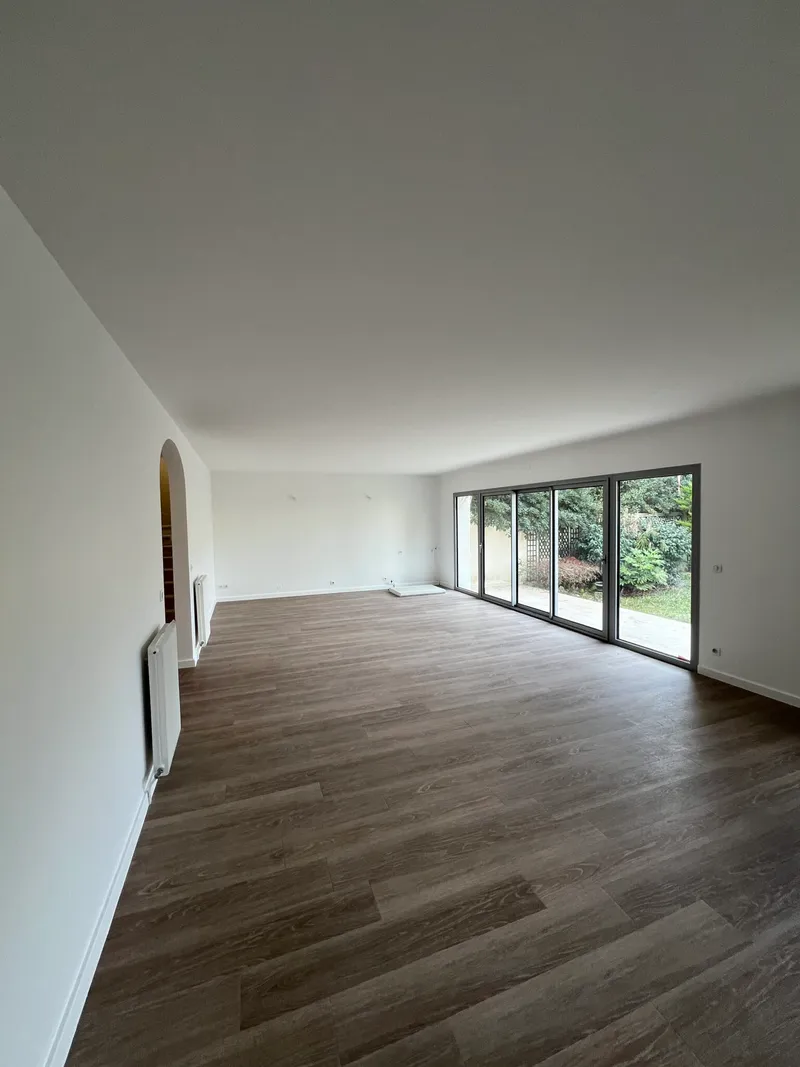 Salon rénové avec parquet brun, murs blancs et baies vitrées donnant sur un jardin