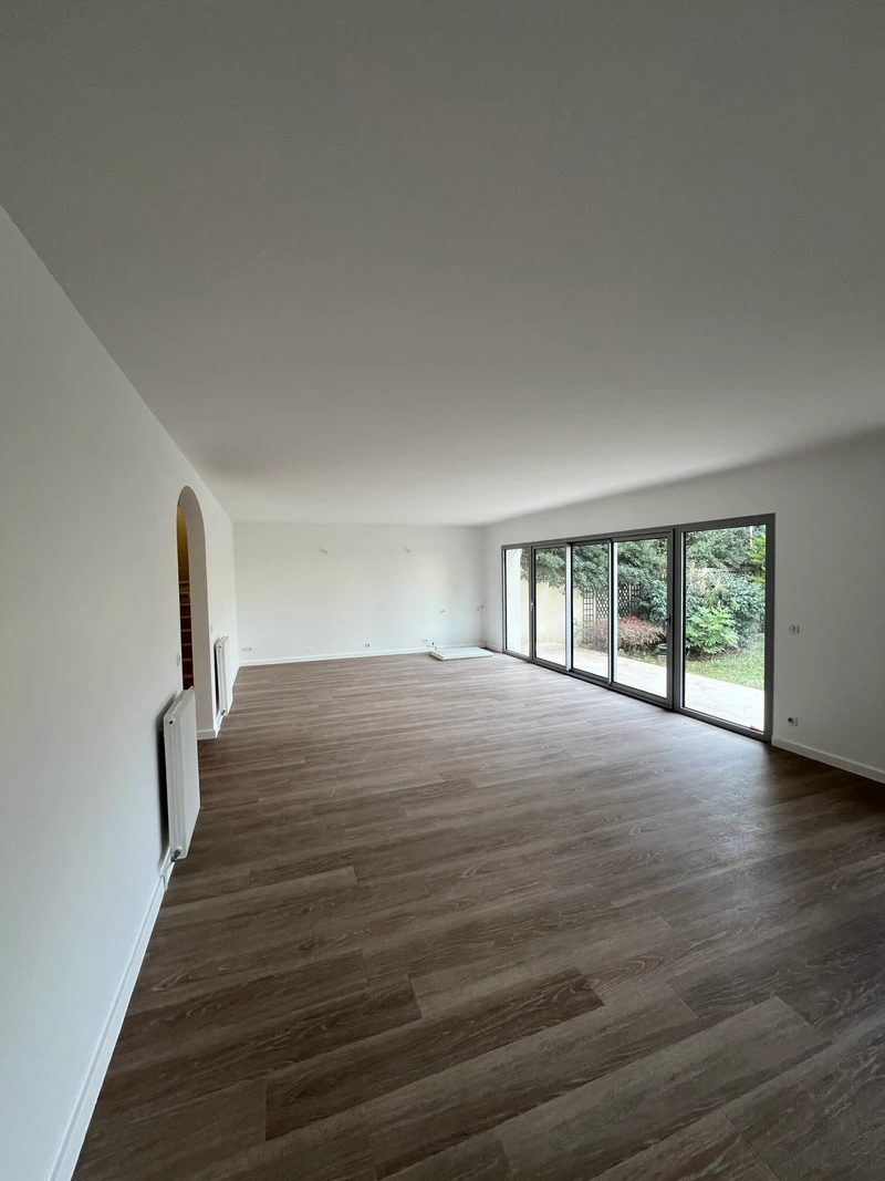 Salon rénové aux murs blancs avec sol brun et larges baies vitrées ouvertes sur un jardin